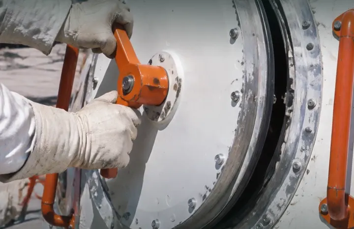 A gloved hand turns the orange handle on a round spacecraft hatch, its metal rim scuffed with use.