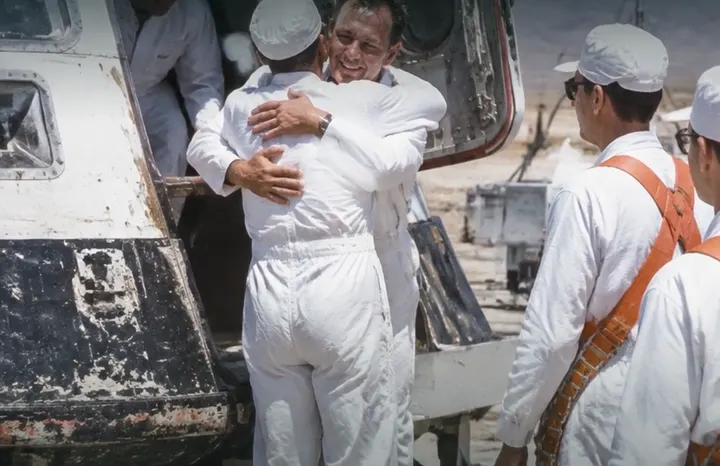 Two people in white flight suits hug in front of an open capsule while a crew member in an orange harness looks on.