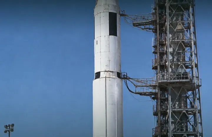 A tall white rocket stands beside its steel launch tower against a deep blue sky.