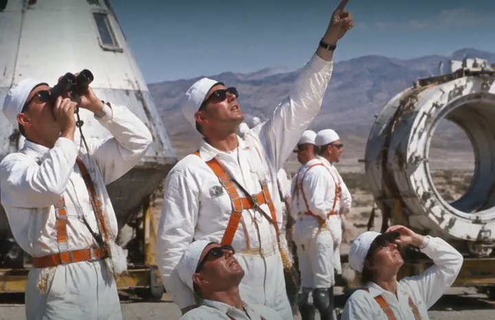 Crew in white suits and orange harnesses watch a launch from the desert, one with binoculars, another pointing skyward.